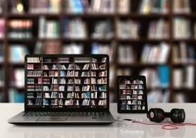 ein PC, ein Tablet, eine Powerbank und Kopfhörer sind vor einem Bücherregal zu sehen